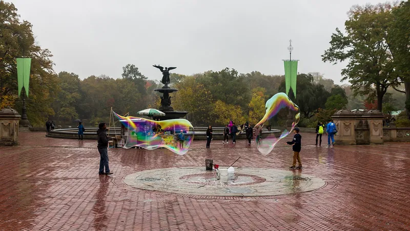 Pompas de jabón gigantes en una plaza junto a una fuente, con gente paseando a lo lejos
