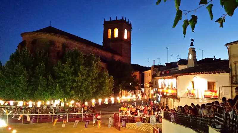 Plaza de un pueblo de la Alcarria en fiestas patronales al anochecer, con la torre de la iglesia iluminada, guirnaldas de bombillas y vecinos en las talanqueras