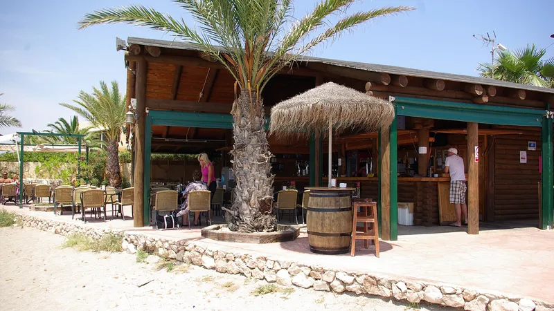Chiringuito de madera con tejado de cañizo, palmeras y mesas al sol junto a la playa
